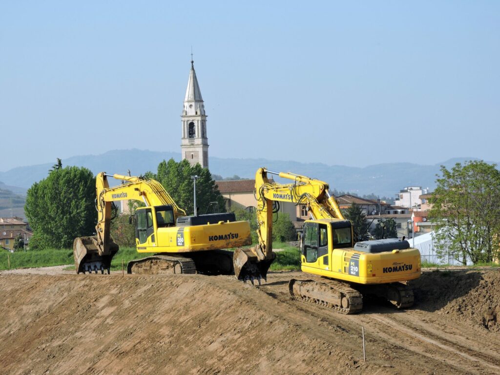 Deux pelles hydrauliques sur chenilles terrassent un terrain. Au loin, un village avec le clocher d'une église.